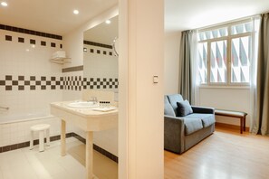 Uma casa de banho moderna com banheira, pia e espelho. Tem uma janela com cortinas e um sofá cinzento na área de estar adjacente.