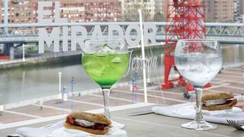 A table with two drinks and sandwiches, a view of a cityscape with a bridge, and the text "El Mirador" on a window.