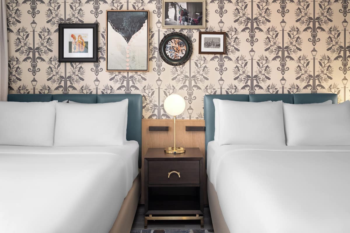 Habitación de hotel con dos camas, una mesita de noche con lámpara y una pared decorada con cuadros.