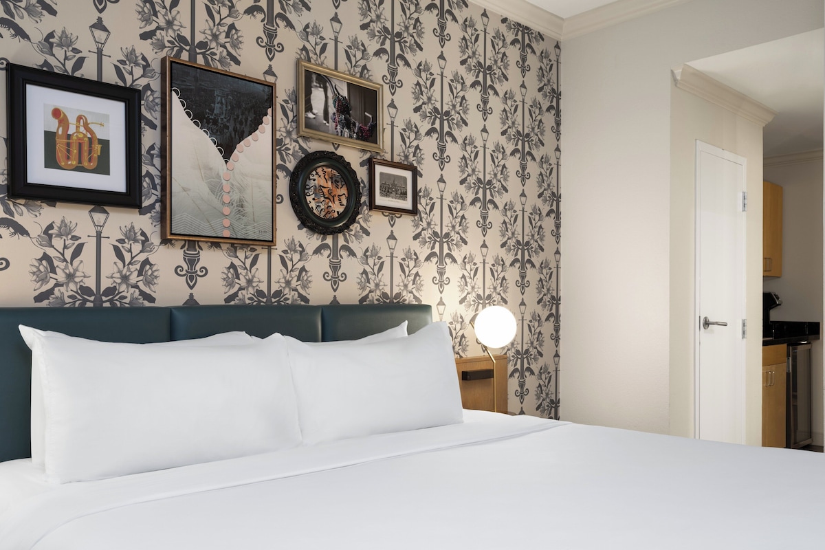A neatly made bed with white linens, a dark green headboard, and a wall adorned with framed artwork and a patterned wallpaper.