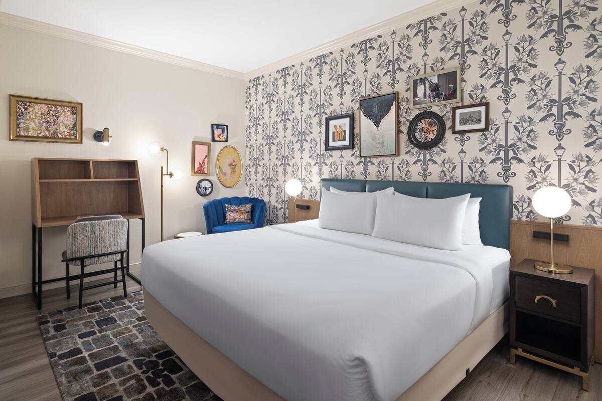 Habitación de hotel ordenada con una cama grande, un escritorio, una silla y una mesita de noche con lámpara.