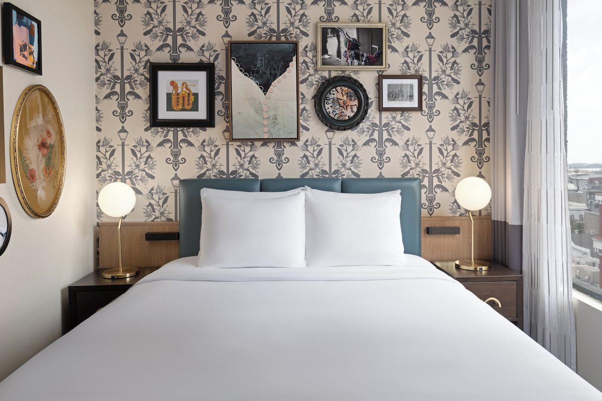 A neatly made bed with white linens, a teal headboard, and two bedside lamps. The room features a patterned wallpaper with framed artwork on the walls.