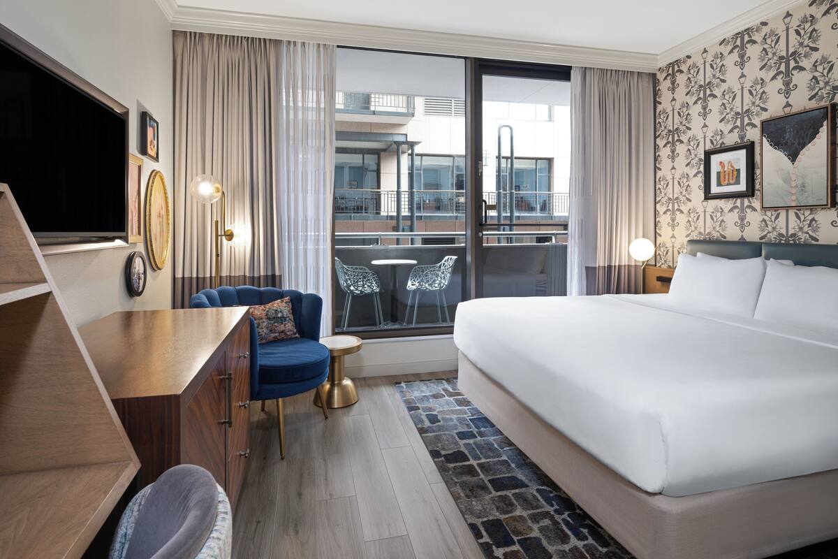 Habitación de hotel con cama, escritorio, silla, televisor y balcón con vista a la ciudad.