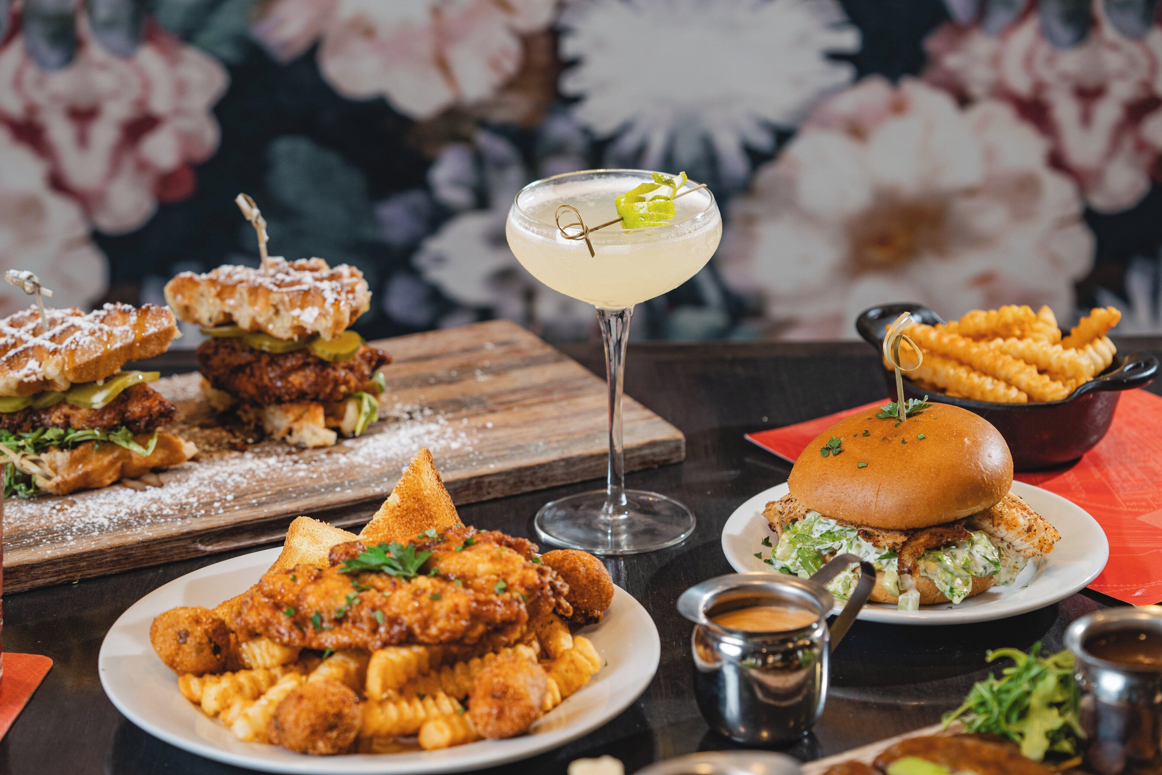 Una mesa con diferentes platos, incluyendo pollo frito, pasta y sándwiches, junto con un cóctel.