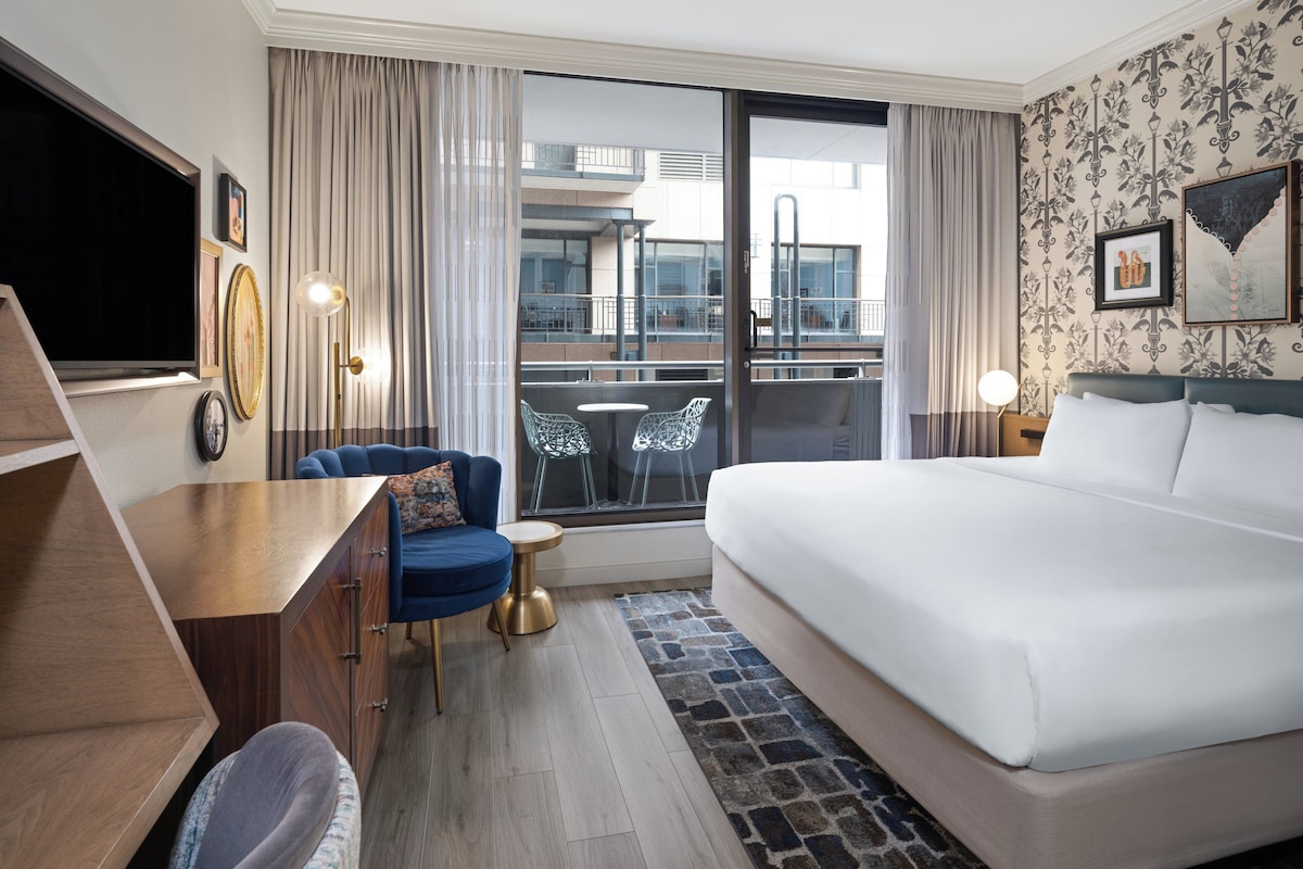 A hotel room with a bed, a desk, a chair, a TV, and a balcony with a view of the city.