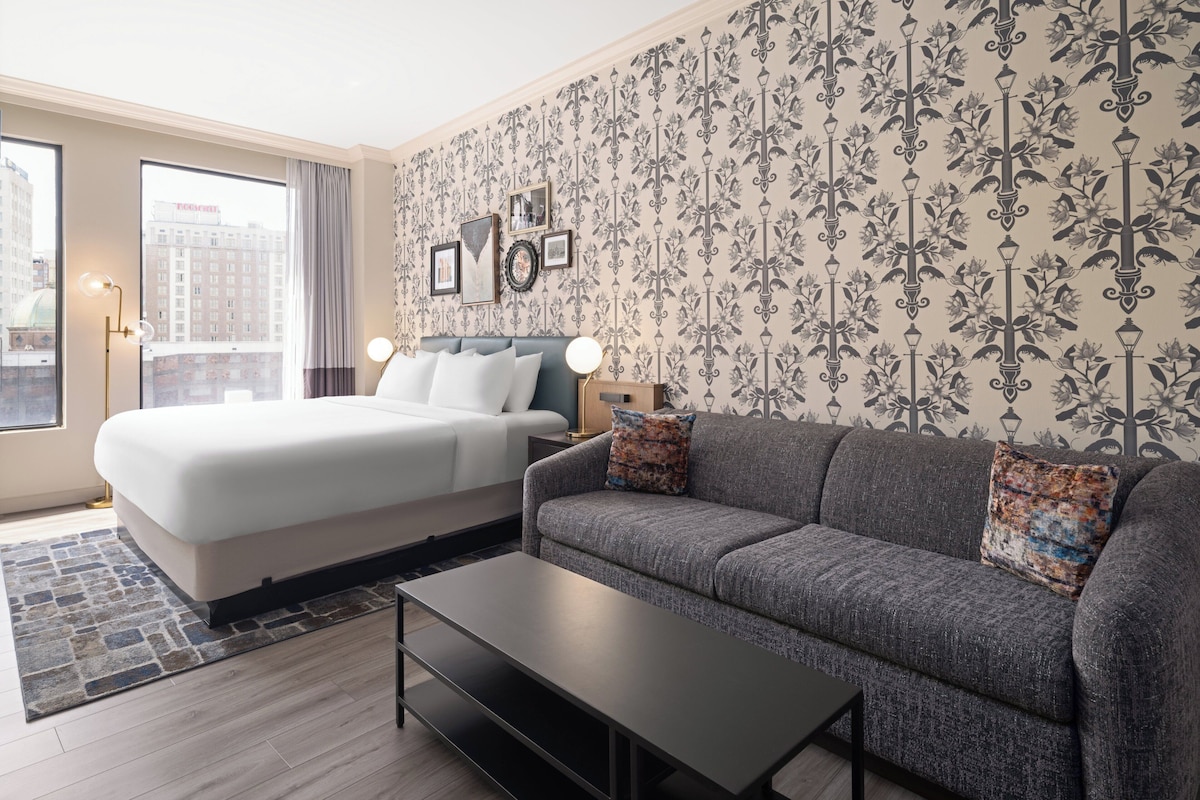 A modern hotel room with a large bed, a grey sofa, a coffee table, and a patterned wallpaper.