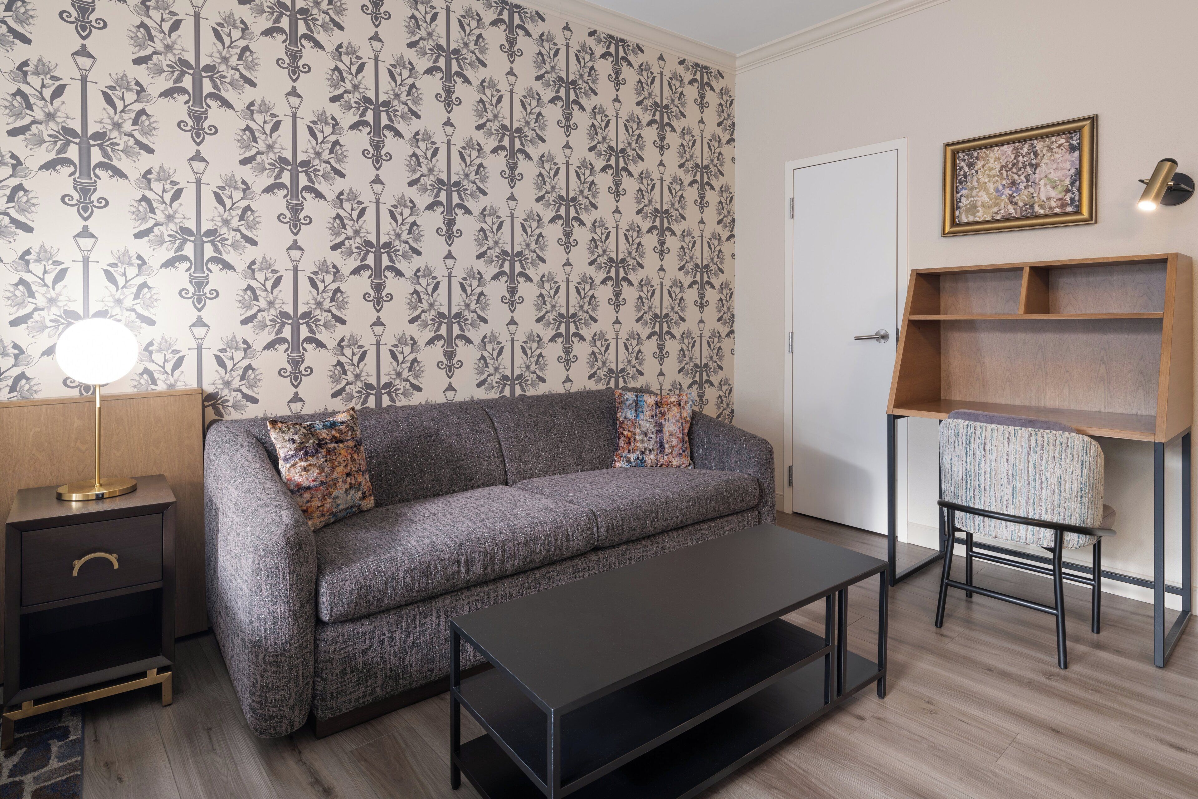 Een woonkamer met een grijze bank, een zwarte salontafel, een bijzettafeltje met een lamp, behang met een patroon en een houten boekenkast.