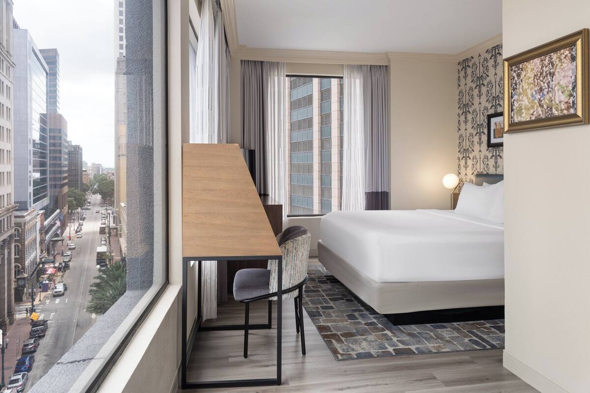 Habitación de hotel con cama, escritorio, silla, ventana con cortinas, vista a la ciudad y un cuadro enmarcado en la pared.