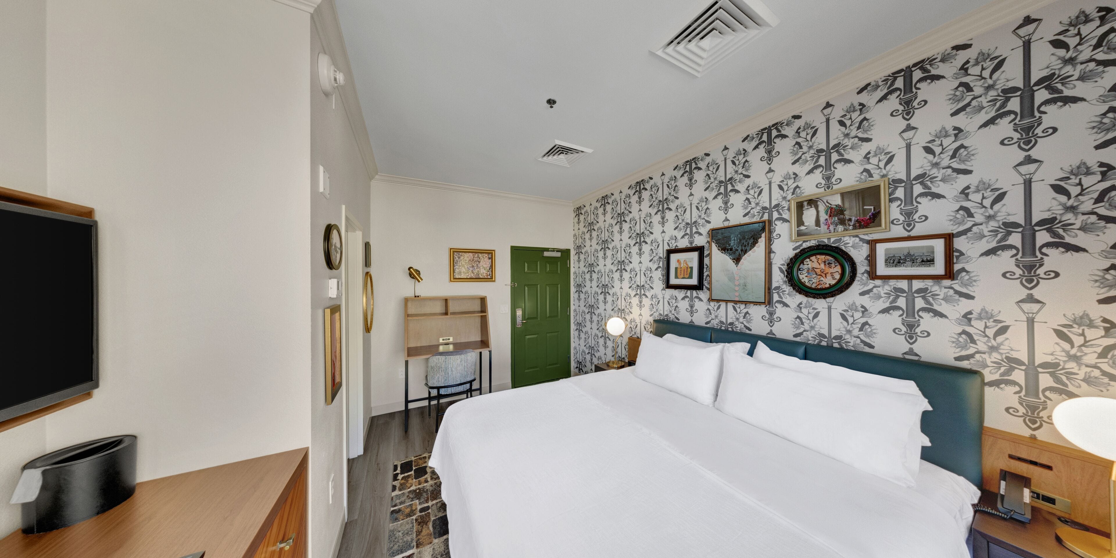 A hotel room with a large bed, a desk with a chair, a TV, and a green door.