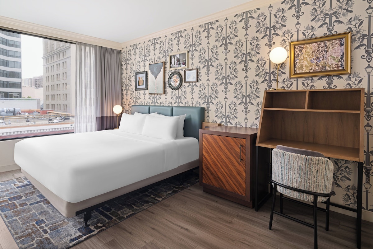 A hotel room with a large bed, a wooden nightstand, a chair, a desk, and a city view through the window.