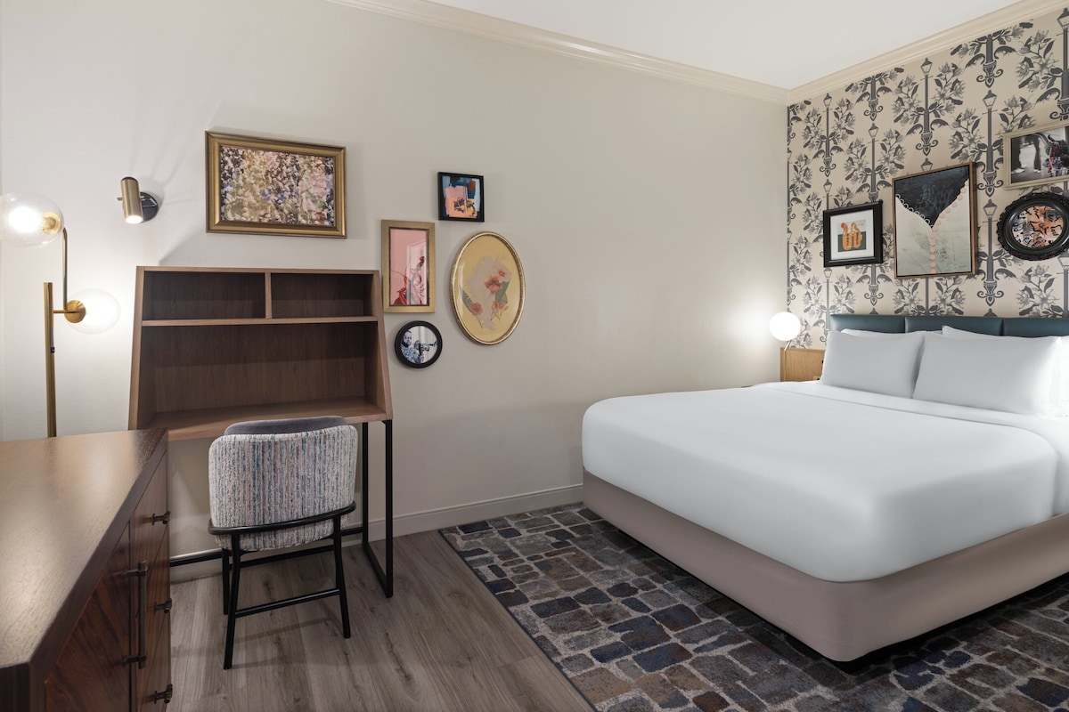 A modern bedroom with a bed, a desk, and a chair. There are wall-mounted shelves and framed pictures.
