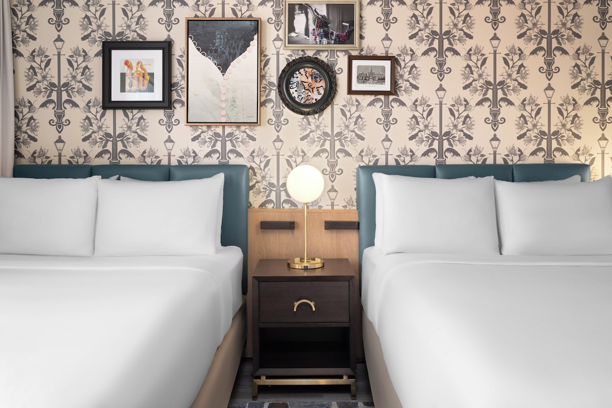 A hotel room with two beds, a nightstand with a lamp, and a wall with framed pictures.