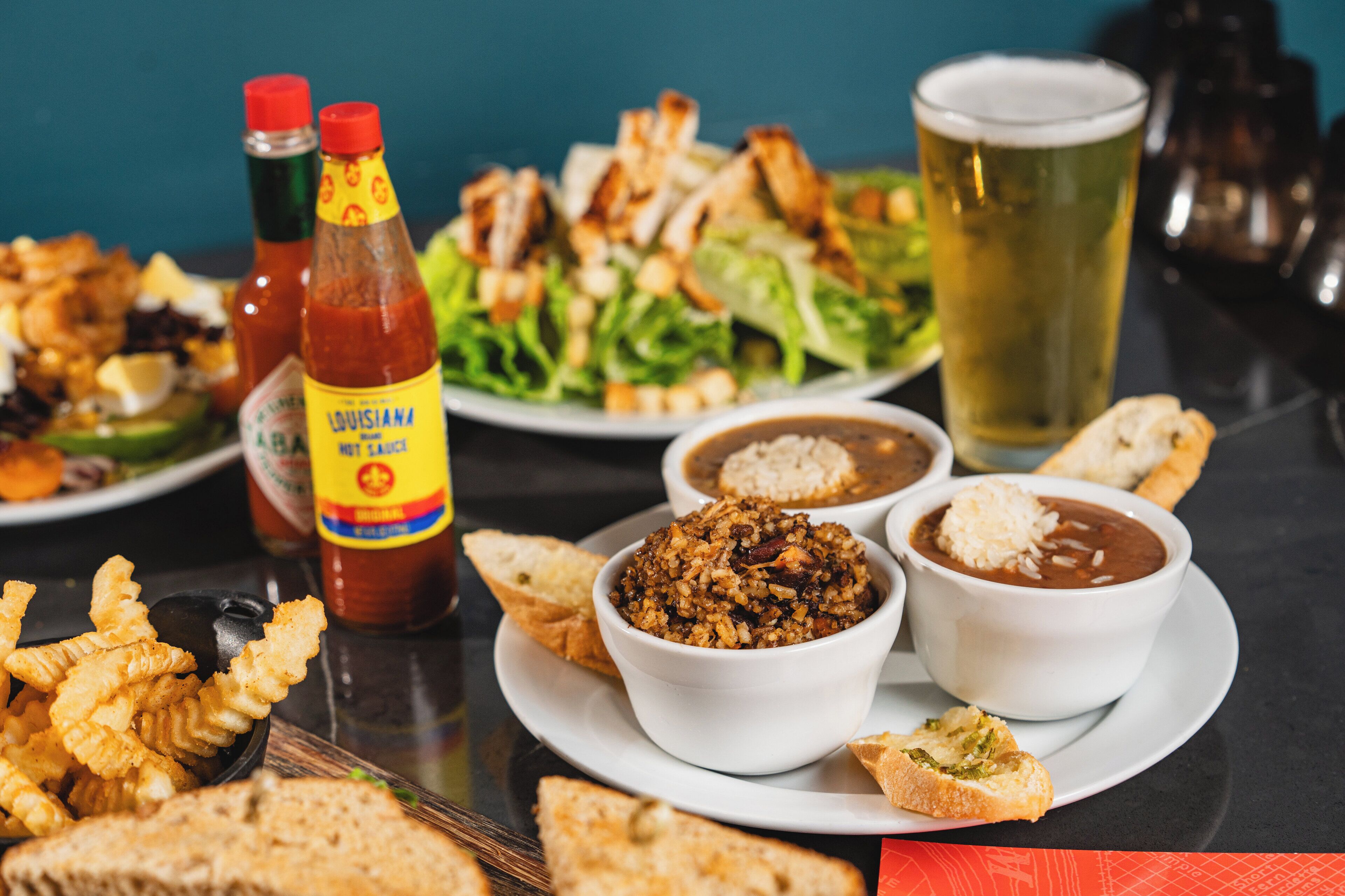 Une table garnie de divers plats, notamment une salade, des bols de chili et des plats frits, ainsi que des bouteilles de condiments et un verre de bière.