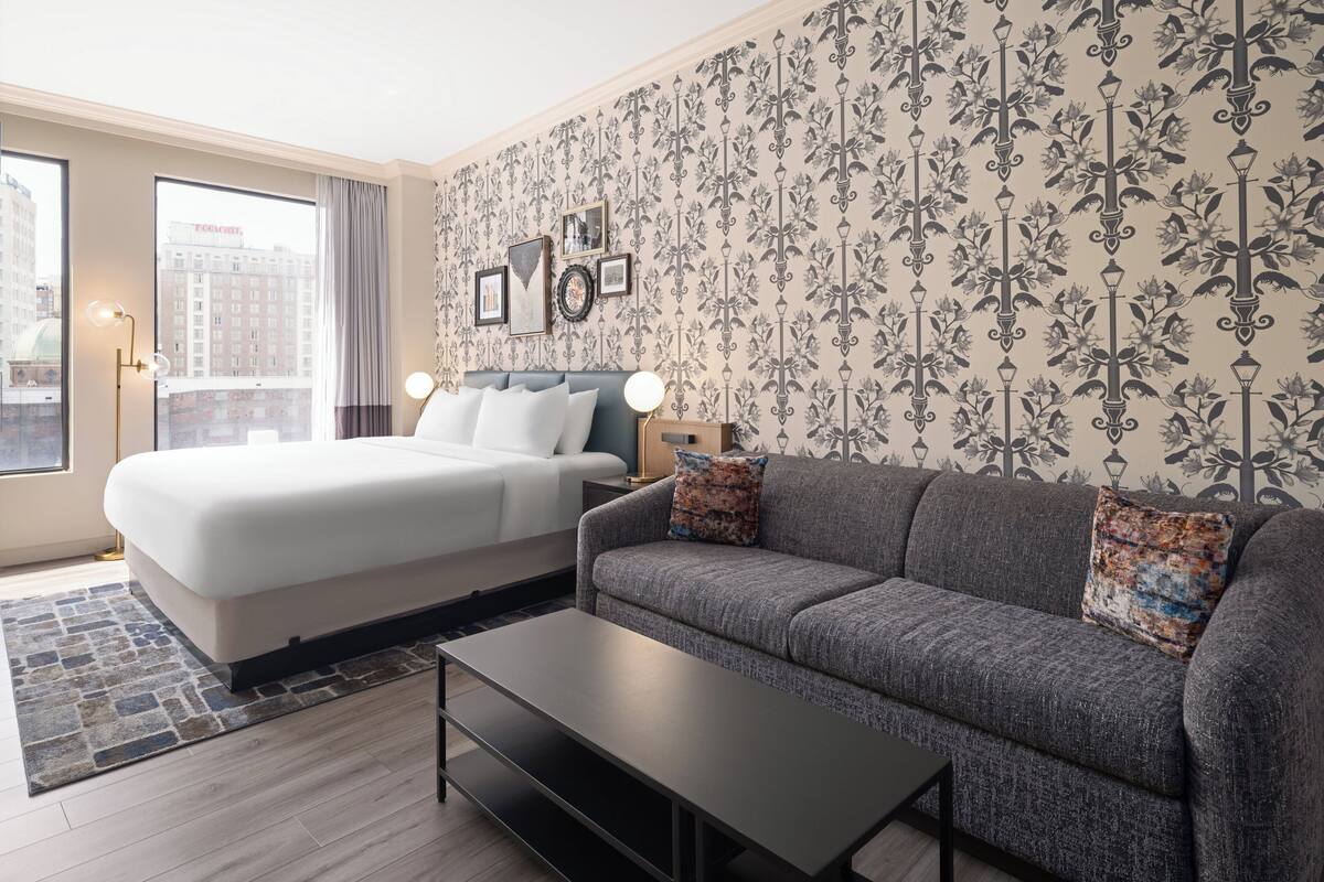 Una habitación de hotel moderna con una cama grande, un sofá gris, una mesita de centro y un papel tapiz estampado.