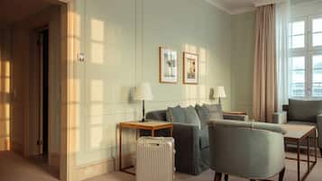 Quarto de hotel com sofá, poltronas, uma mesinha, luminária, mala e quadros nas paredes.