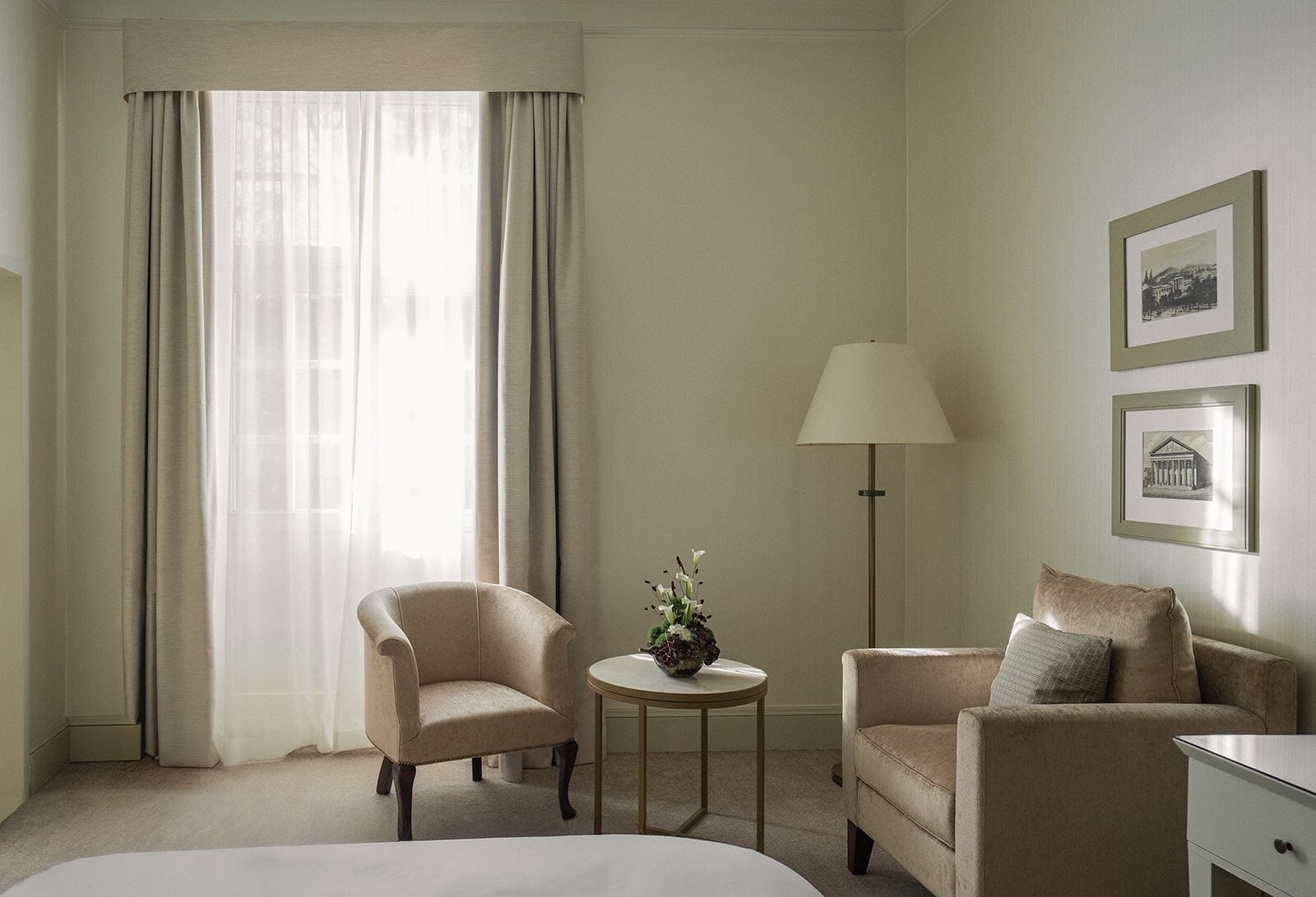 Ein Hotelzimmer mit einem Bett, zwei Sesseln, einem kleinen runden Tisch mit einer Vase, einer Stehlampe und gerahmten Bildern an der Wand.