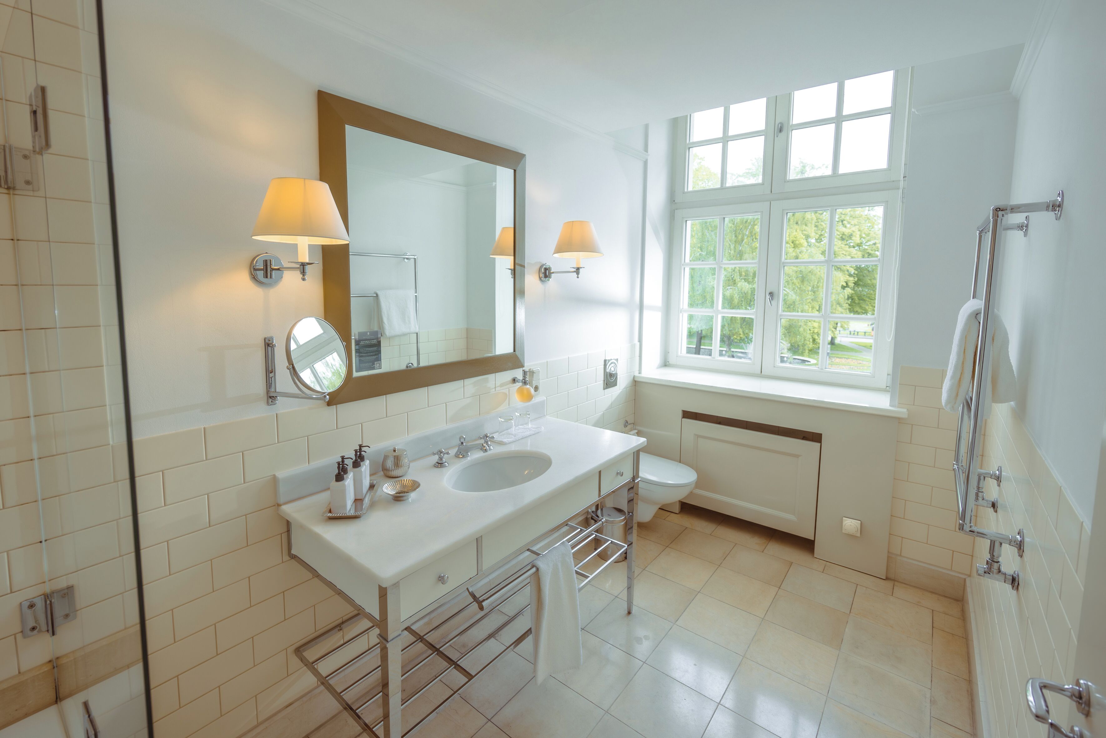 A bathroom with a double sink, a large mirror, two wall-mounted lamps, a window with a view, and a towel rack.