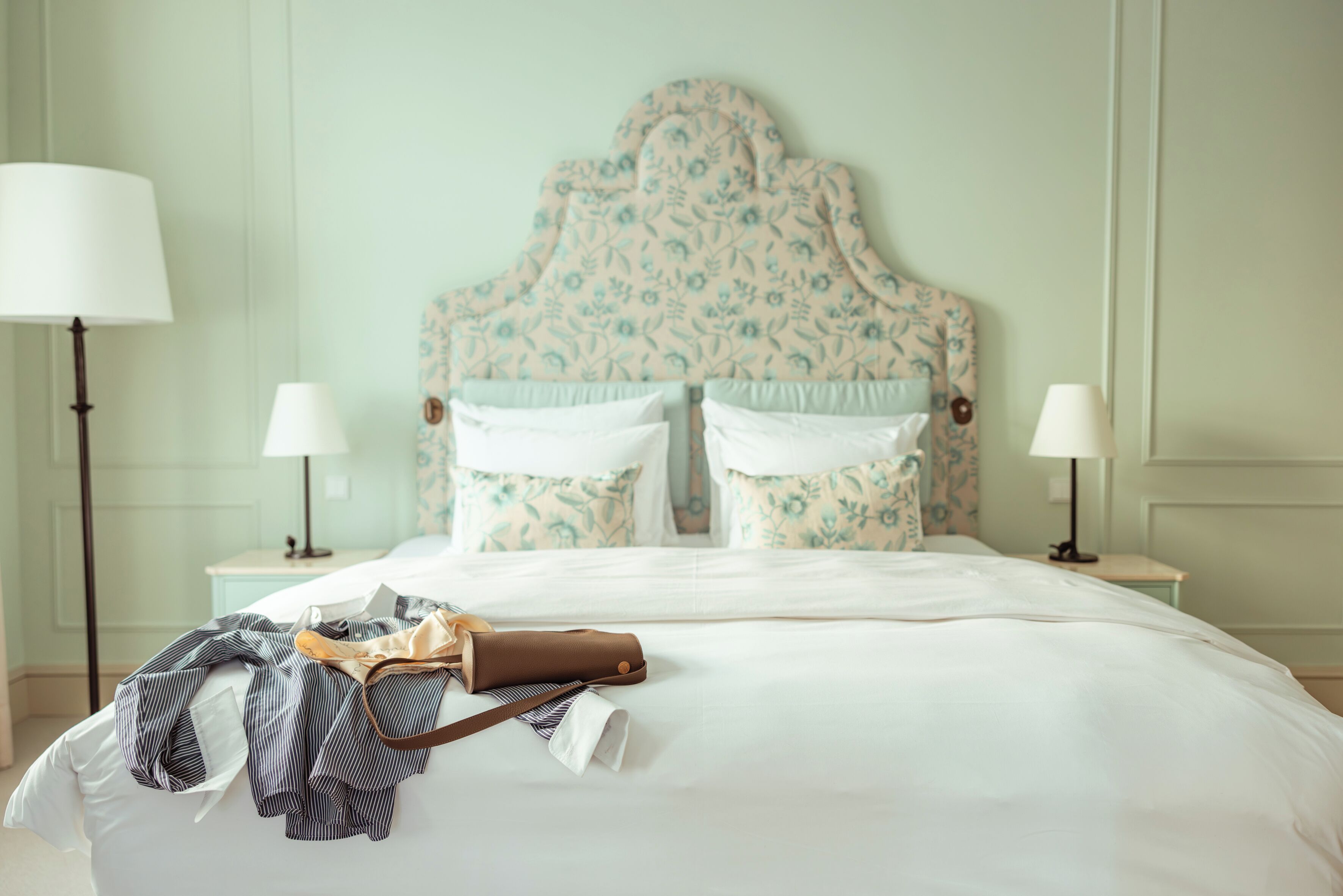 Un lit bien fait, avec une tête de lit à motifs floraux, deux lampes de chevet et un sac en cuir marron posé sur le lit.