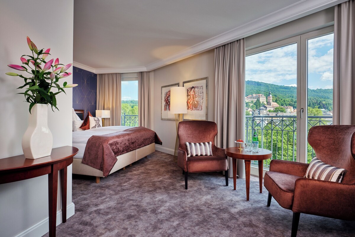 A hotel room with a bed, two armchairs, a small table, a vase with flowers, and a large window with a view.