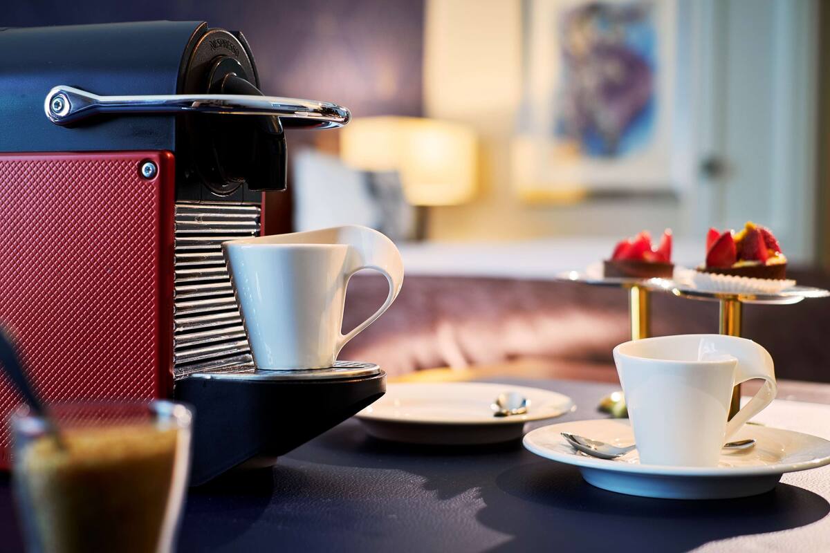 A coffee machine, a white cup, a saucer, a plate with a dessert, and a spoon on a table.