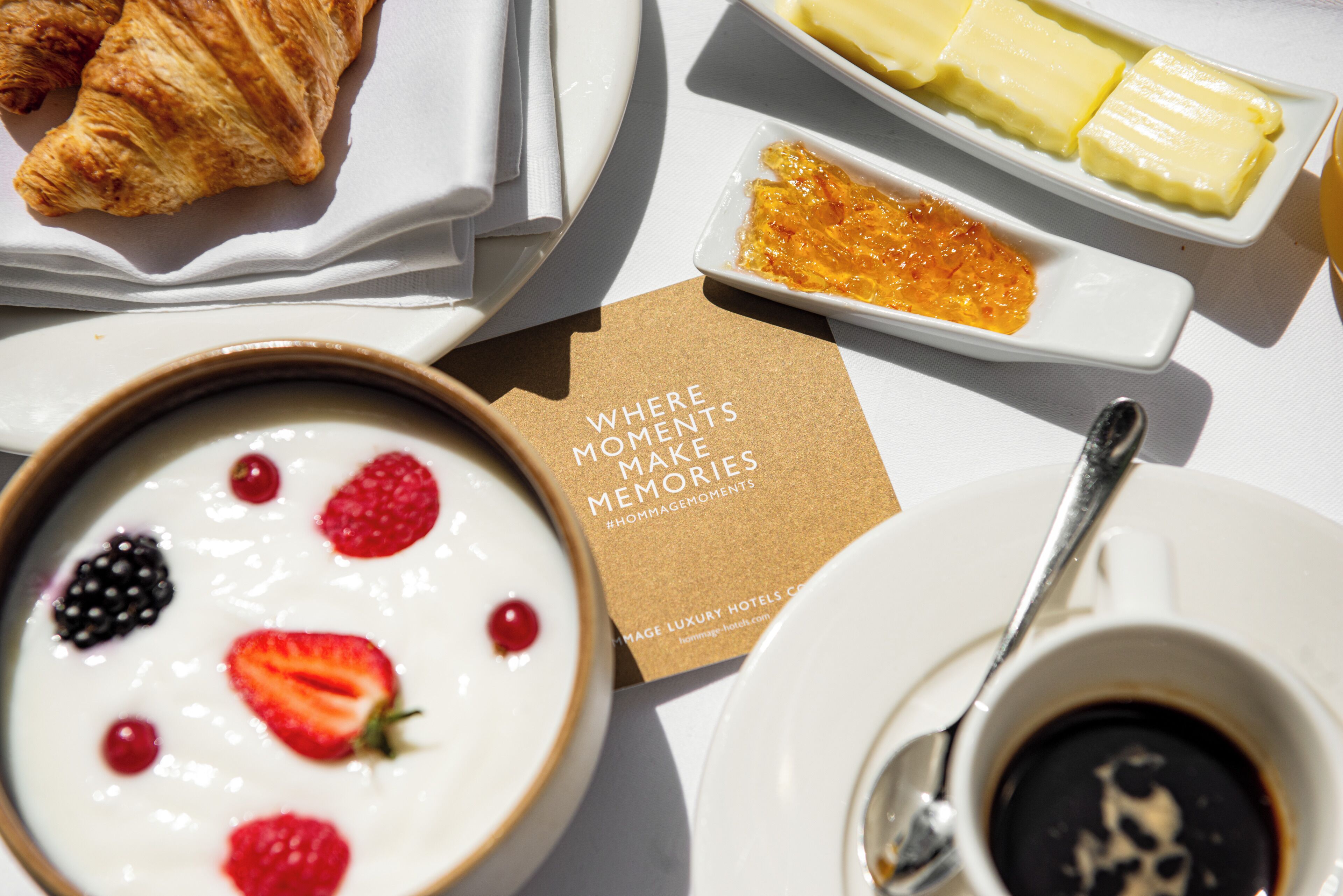 Uma mesa de café da manhã com um croissant, iogurte com frutas vermelhas, um pires de geleia, um guardanapo, uma colher e uma xícara de café.