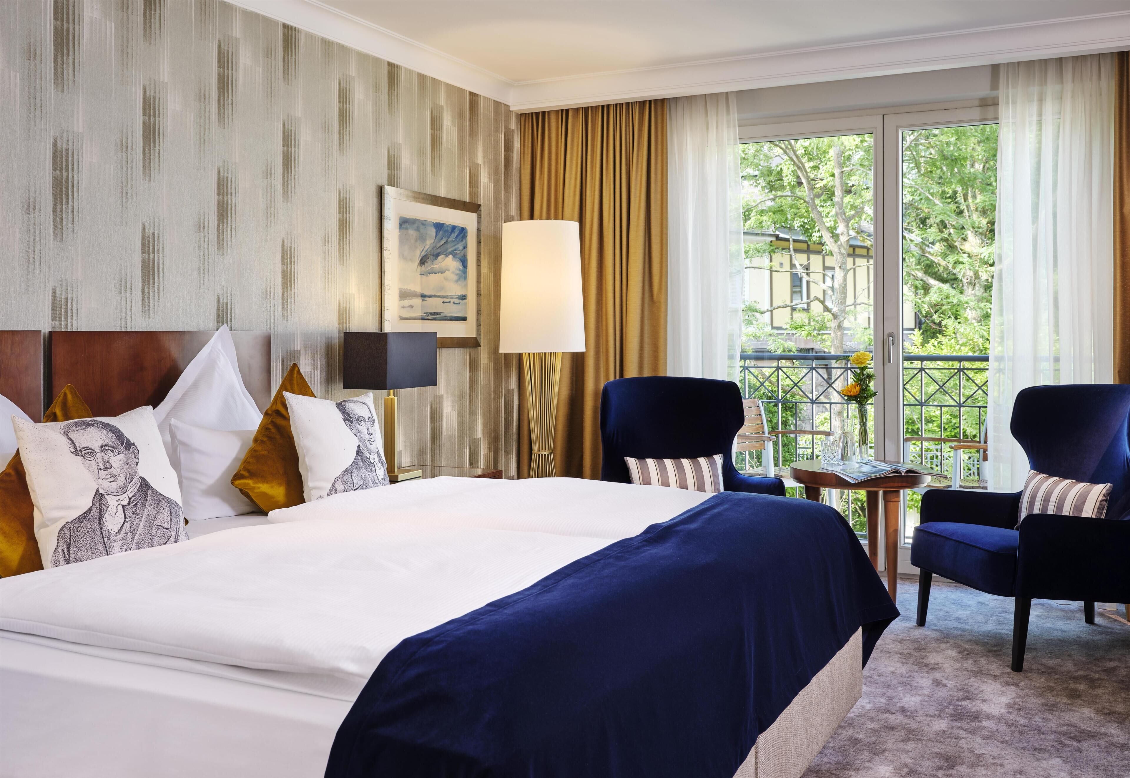 A hotel room with a large bed, two armchairs, a small table, a painting on the wall, and a view of greenery outside.