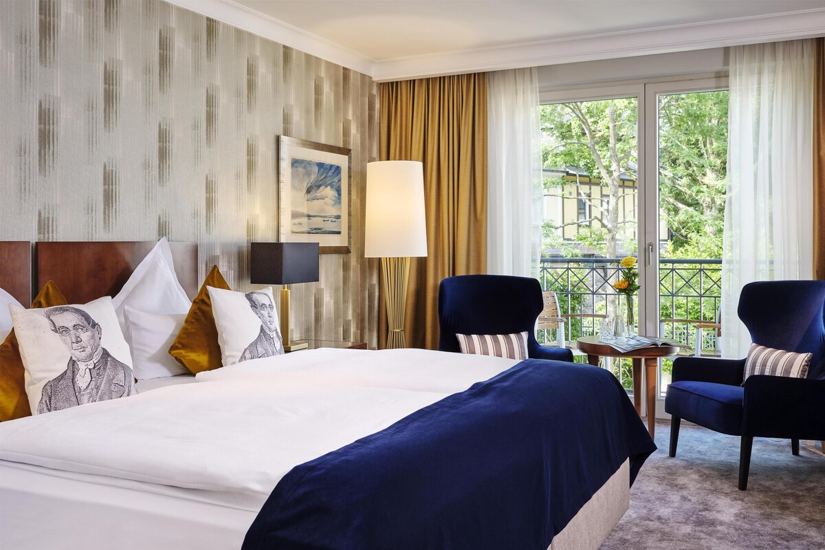 A hotel room with a large bed, two armchairs, a small table, a painting on the wall, and a view of greenery outside.