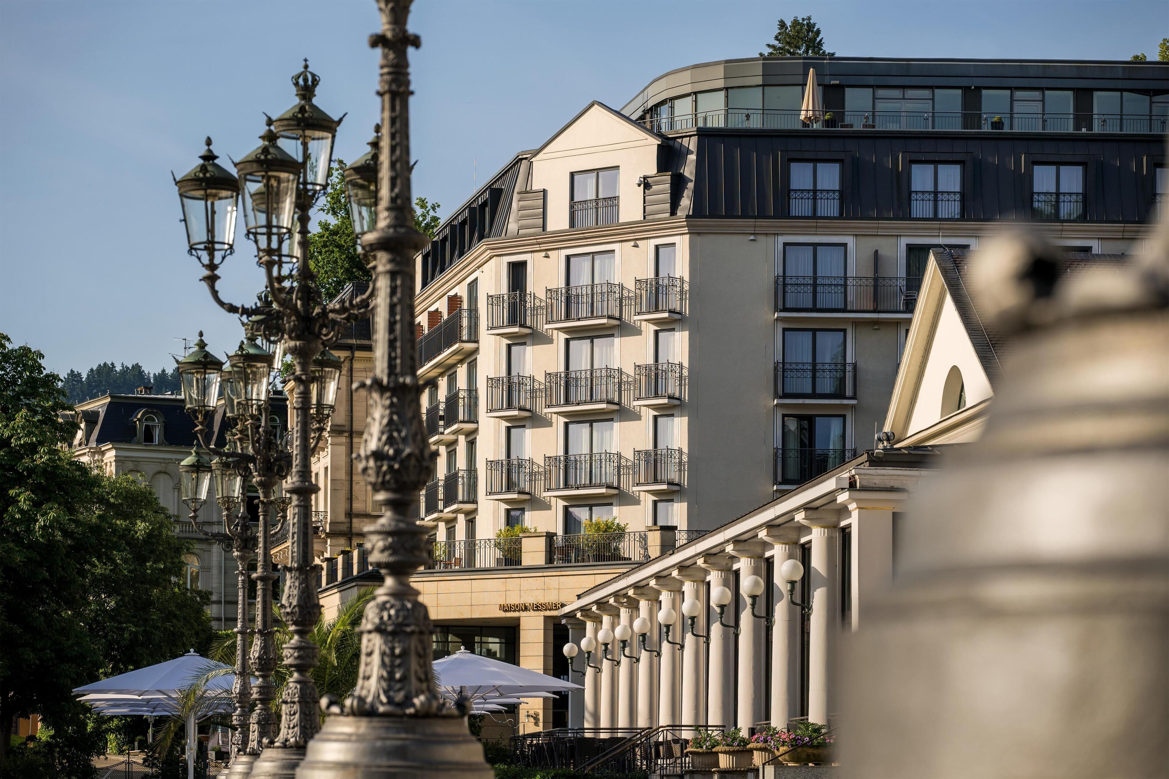 Un edificio de varios pisos con balcones, una zona de café con mesas al aire libre y farolas decorativas.