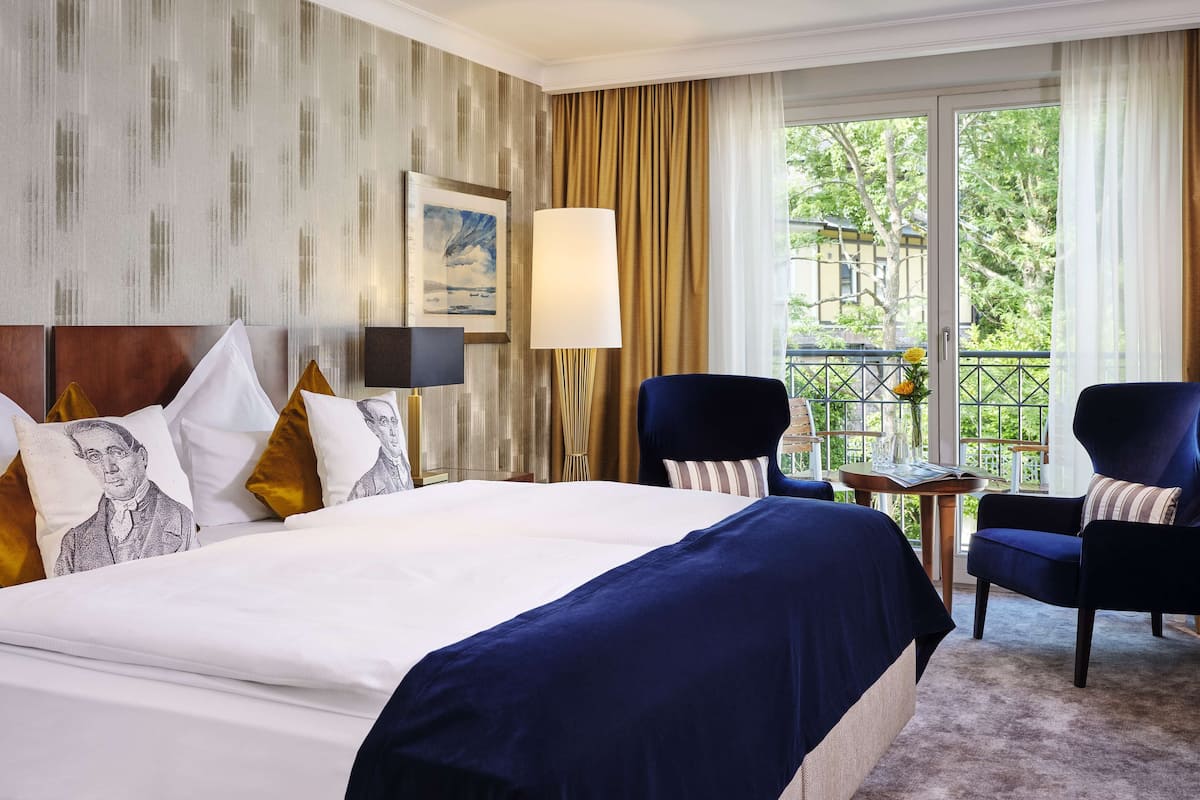 A hotel room with a large bed, two armchairs, a small table, and a view of greenery outside.