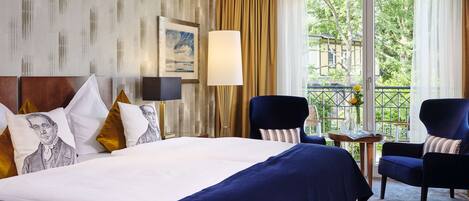A hotel room with a large bed, two armchairs, a small table, and a view of greenery outside.