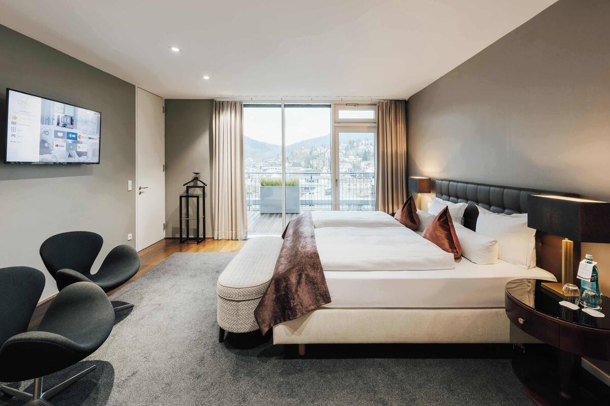 A modern hotel room with a large bed, a TV on the wall, and a balcony with a city view.
