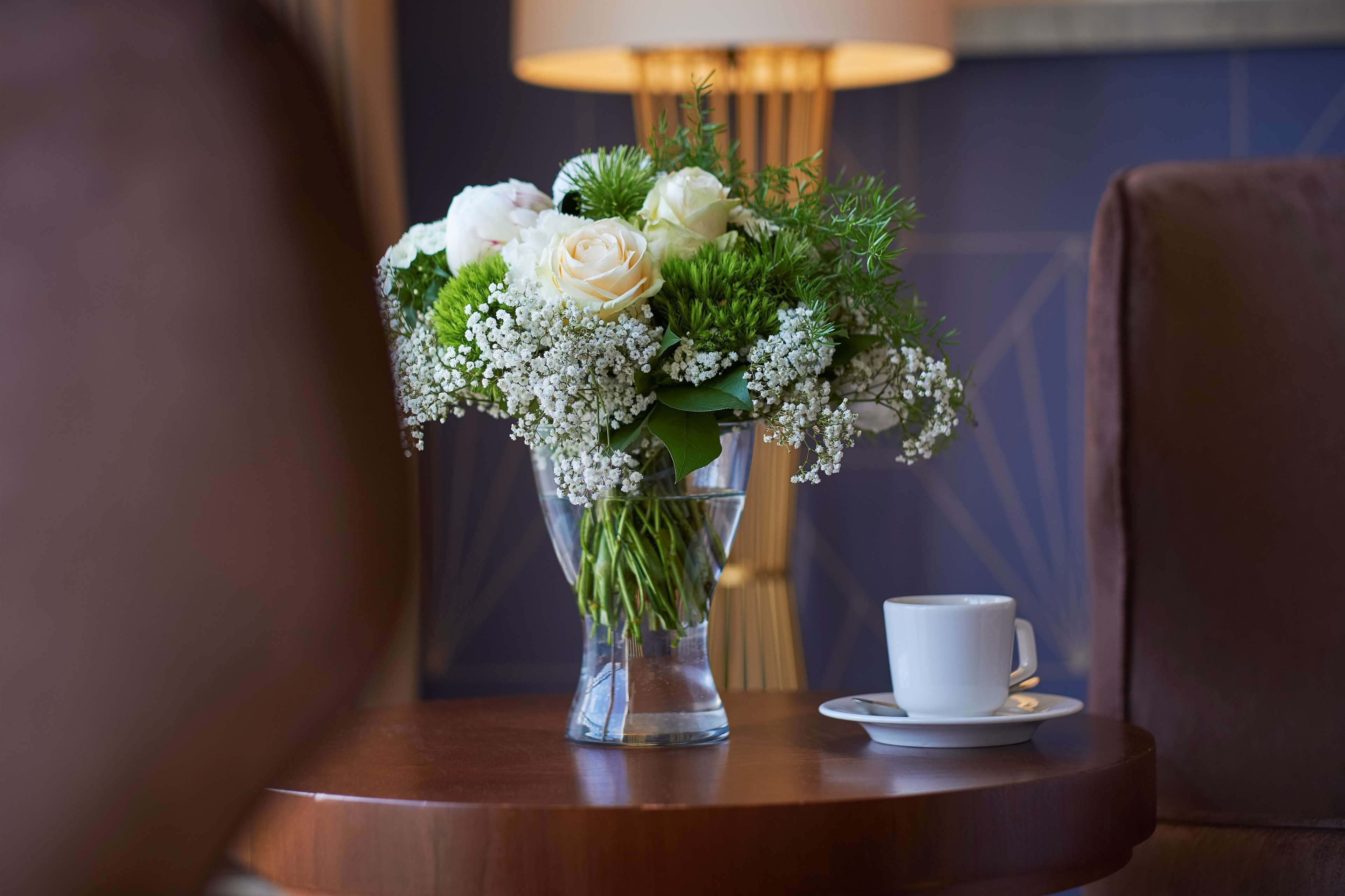 A clear glass vase with white flowers and greenery on a wooden table, accompanied by a white cup and saucer.