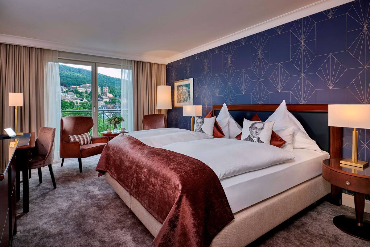 A hotel room with a large bed, two bedside tables, a desk, a chair, and a view of a cityscape through large windows.
