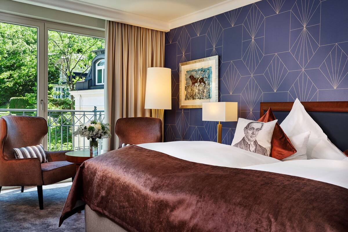 A hotel room with a large bed, two armchairs, a small table with a vase of flowers, and a view of a building through large windows.