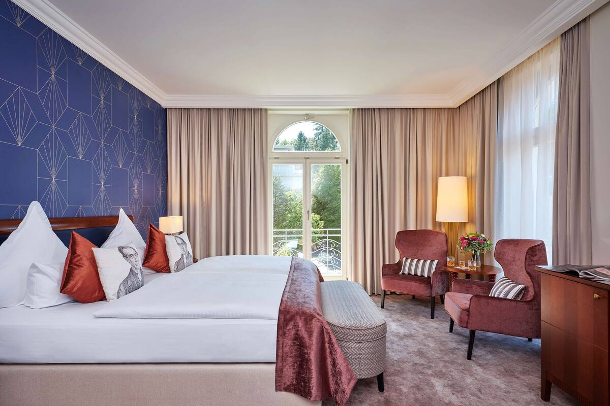 A hotel room with a large bed, two chairs, a desk, and a view of greenery through the window.