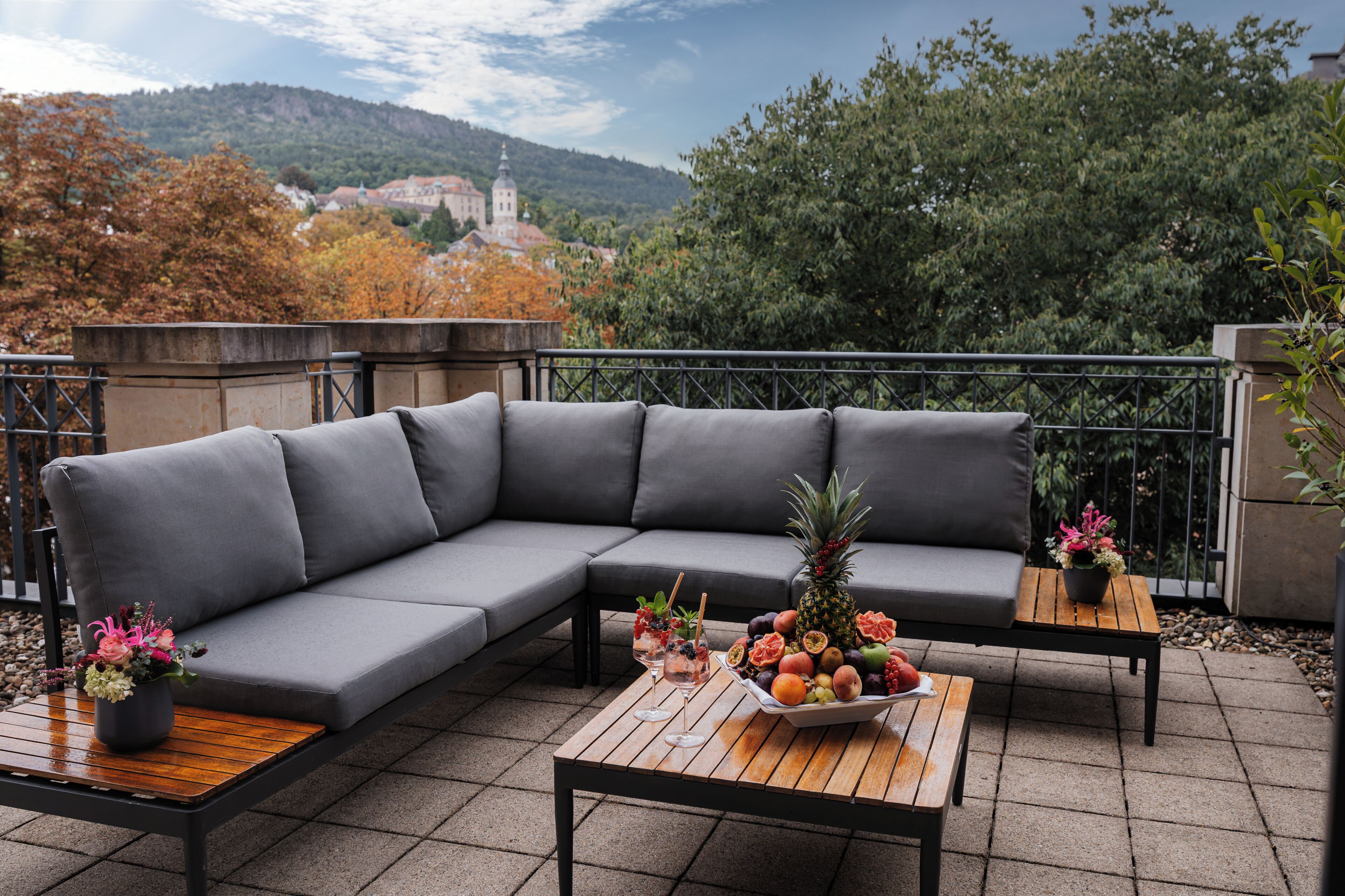 A balcony with a grey sofa, a wooden table with a fruit bowl and drinks, and a flower pot.