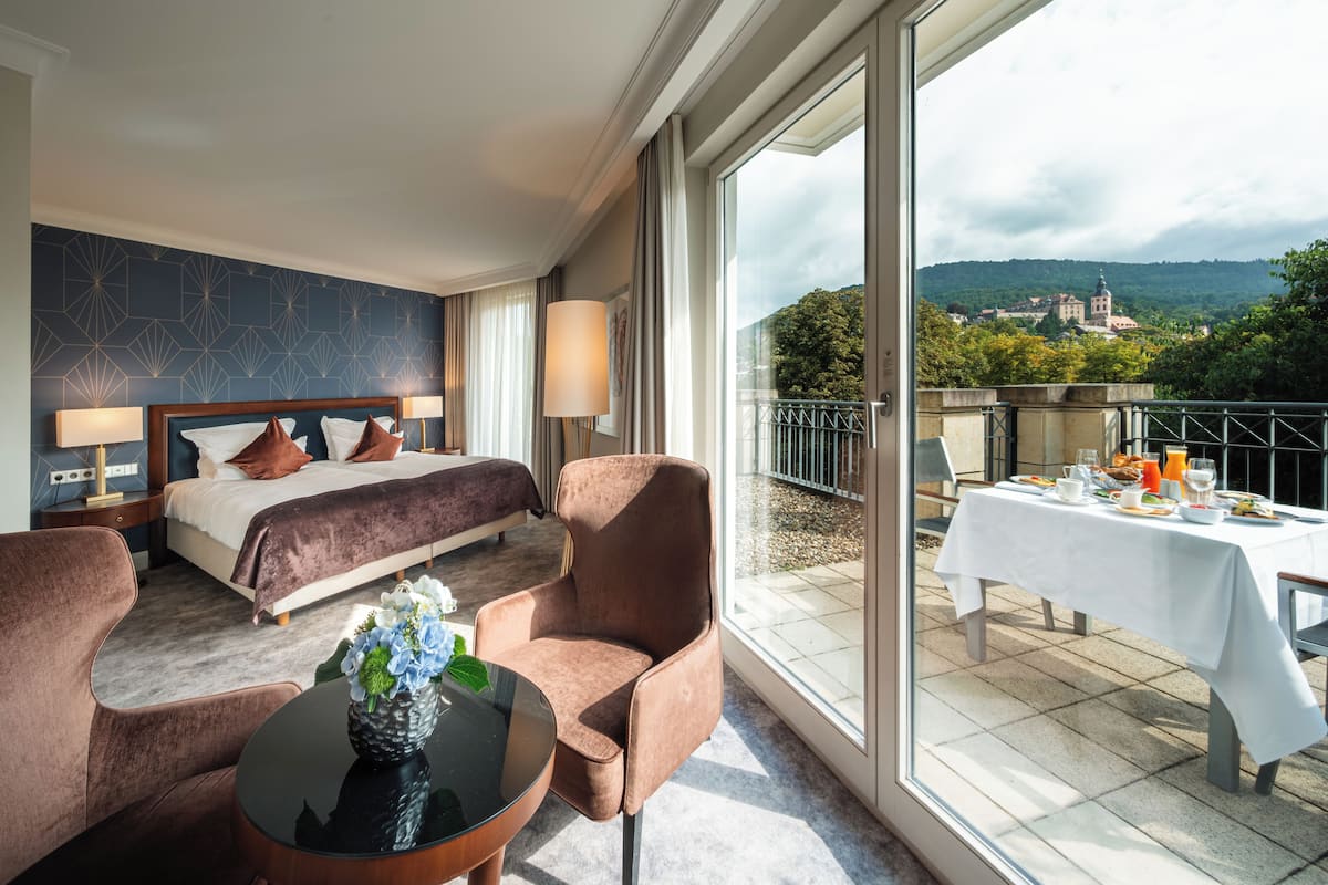 A hotel room with a bed, two chairs, a table, and a balcony with a table set for breakfast.