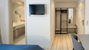 Una habitación de hotel moderna con una cama, un televisor instalado en la pared, un baño con espejo y lavamanos, y una zona de estar con una silla y mesa.