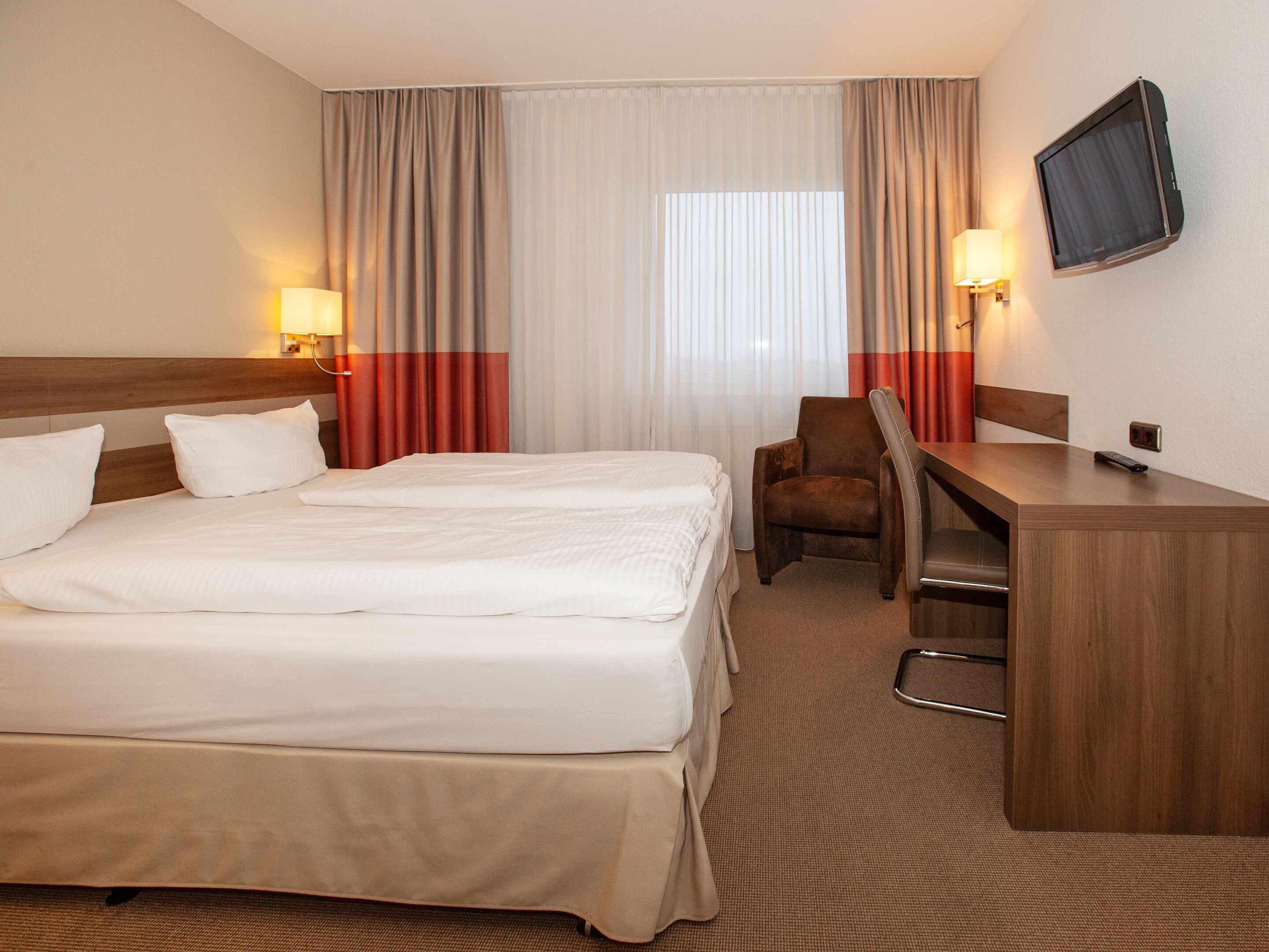 A hotel room with a large bed, a desk with a chair, a television mounted on the wall, and curtains on the windows.