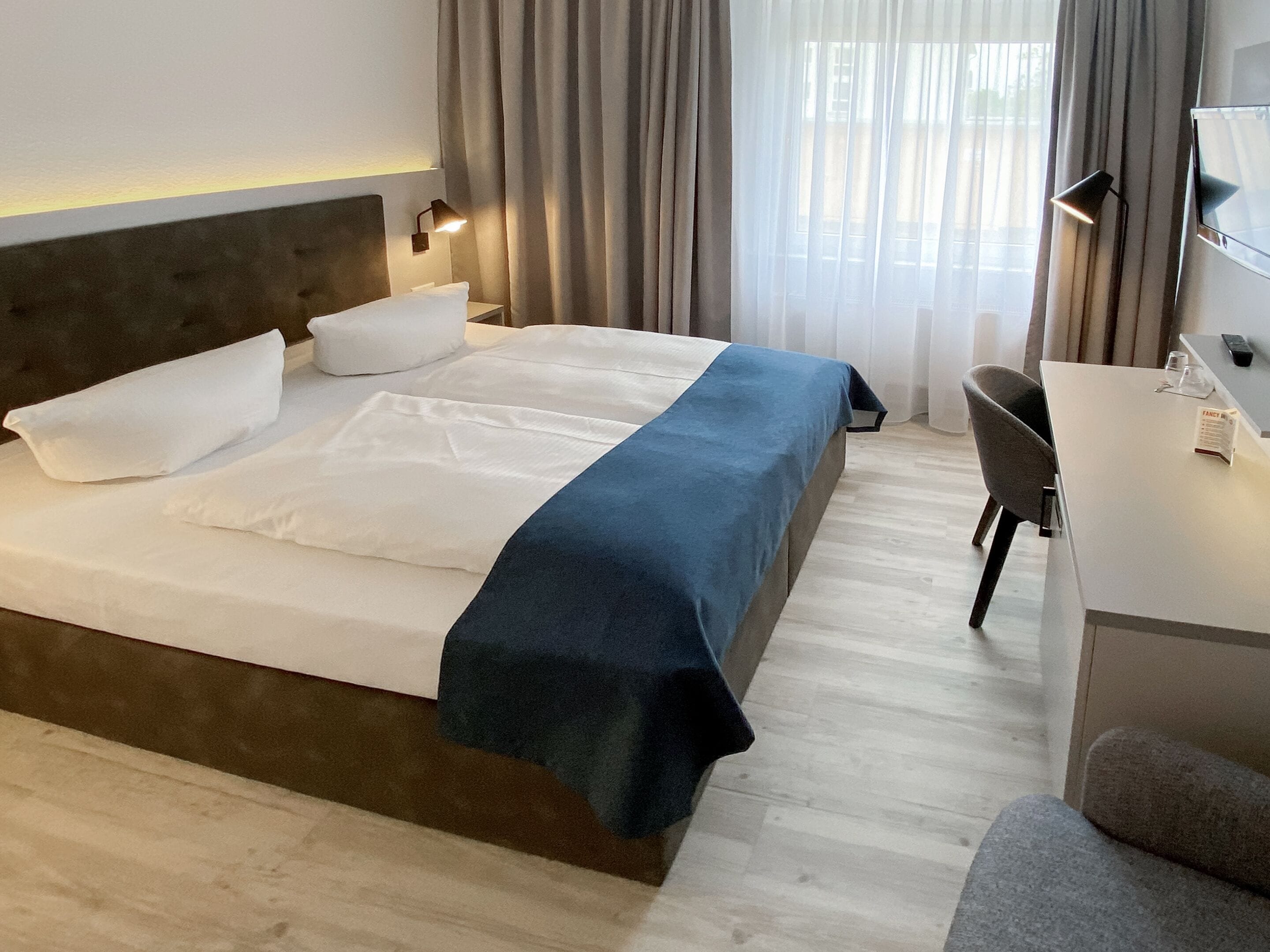 Una cama bien hecha con ropa de cama blanca y azul, una lámpara de noche y un escritorio con una silla.