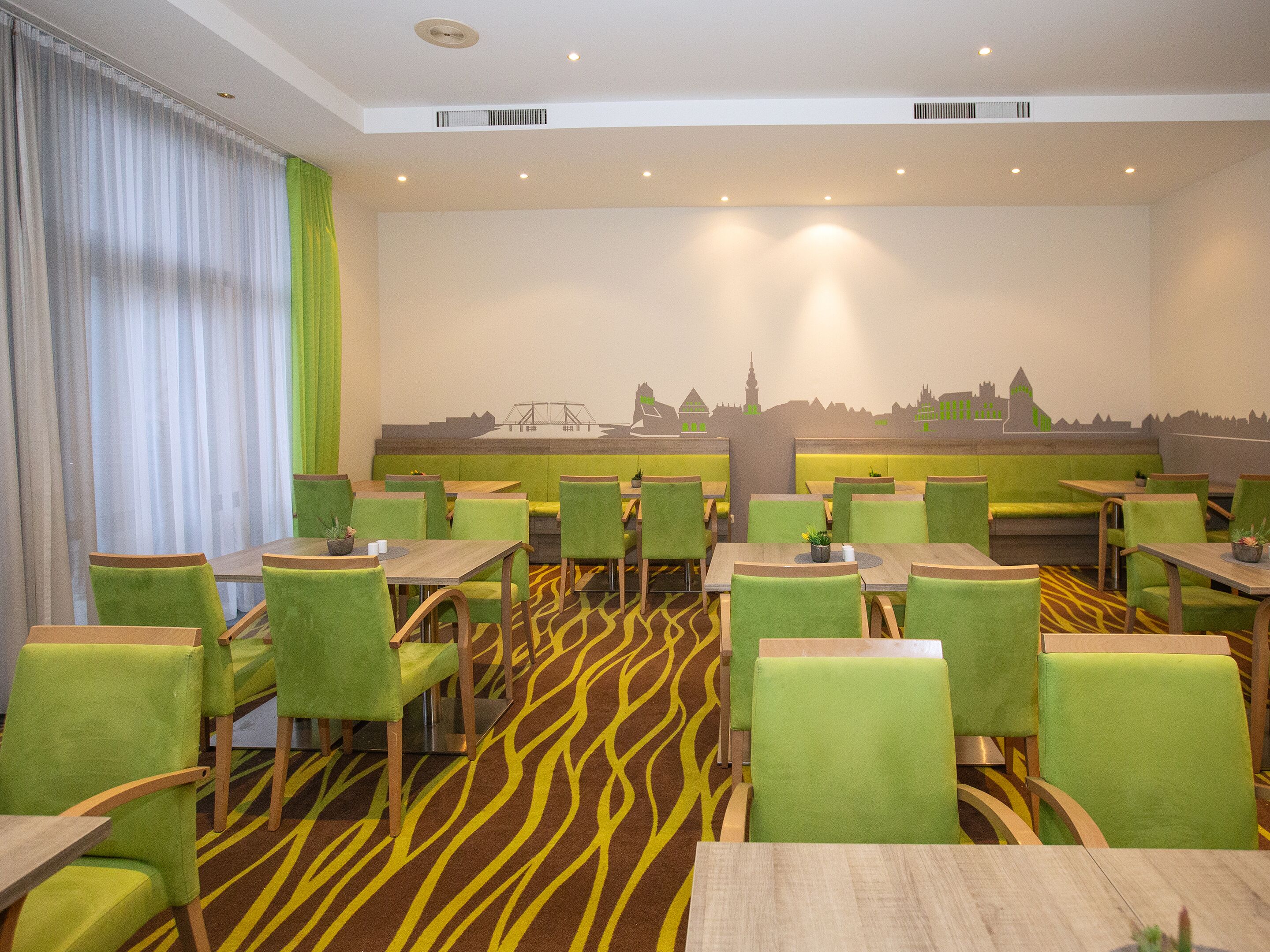A dining area with green upholstered chairs and tables, a patterned carpet, and a mural on the wall depicting a city skyline.