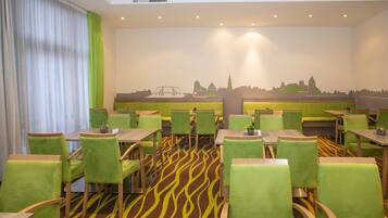 Un comedor con sillas tapizadas de verde y mesas, una alfombra estampada y un mural en la pared que representa el horizonte de una ciudad.