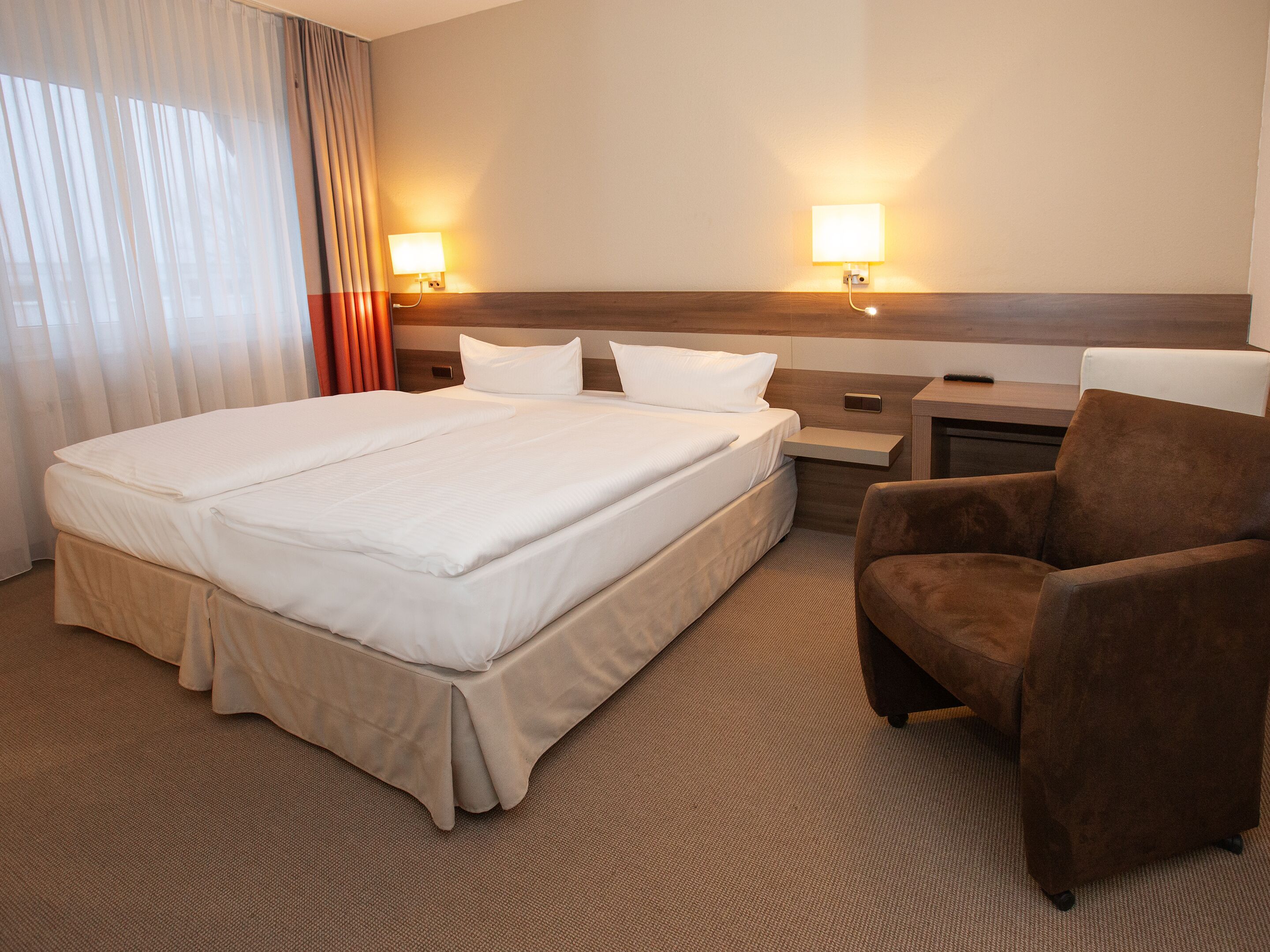 A hotel room with a large bed, two bedside tables, a chair, and two wall-mounted lamps.