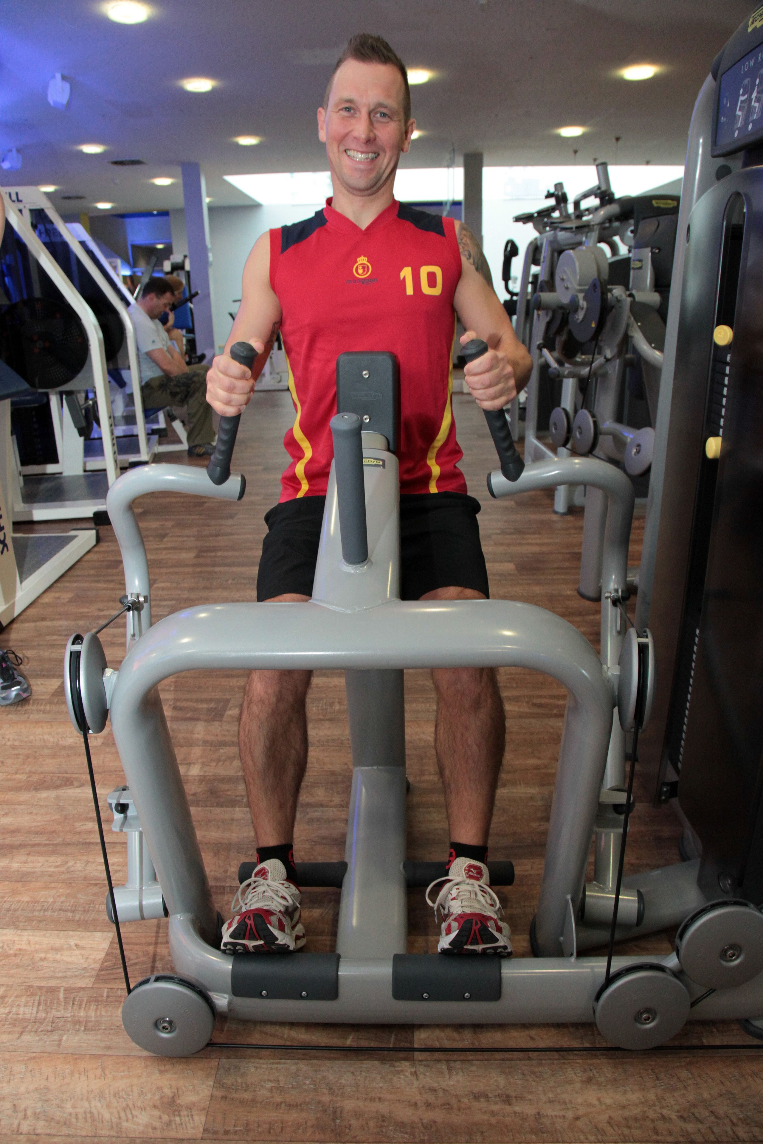 Een man aan het sporten op een hometrainer in een sportschool, met diverse apparaten op de achtergrond.