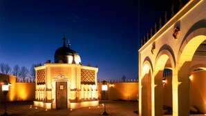 A courtyard with a central building featuring a dome and a minaret, surrounded by an arched structure with illuminated columns.