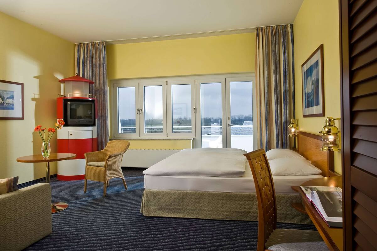 A hotel room with a bed, a small table with a vase, a chair, a television, and a window with a view of the outside.