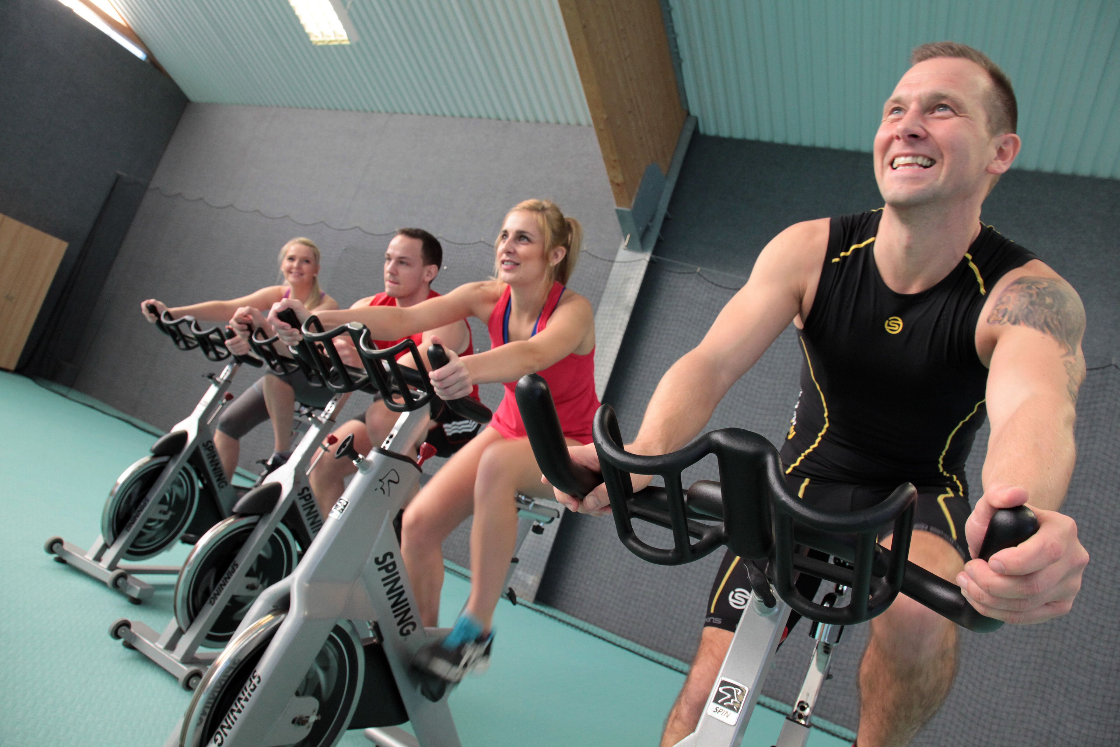 Trois personnes font du vélo d’appartement dans une salle de sport, devant un mur gris et sur un sol bleu.