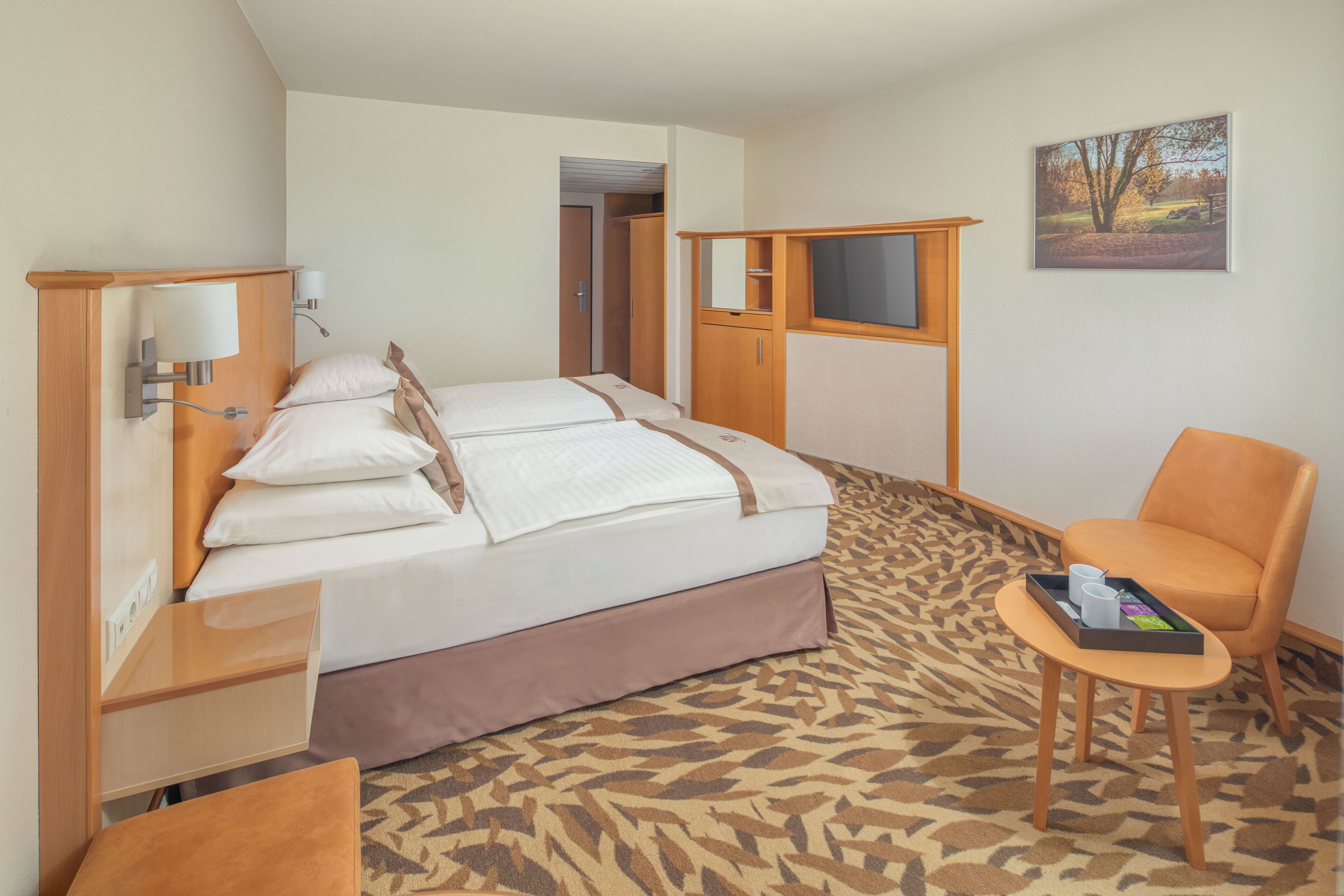 A hotel room with a bed, a chair, a small table, and a painting on the wall.