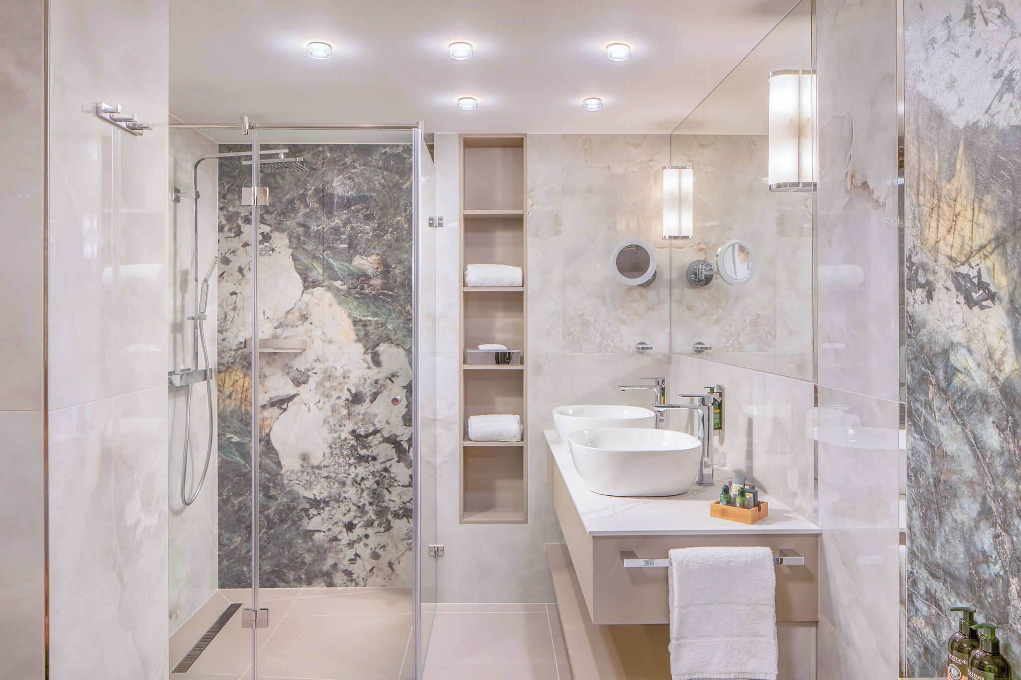 Un baño moderno con ducha de vidrio, paredes de mármol y tocador doble.
