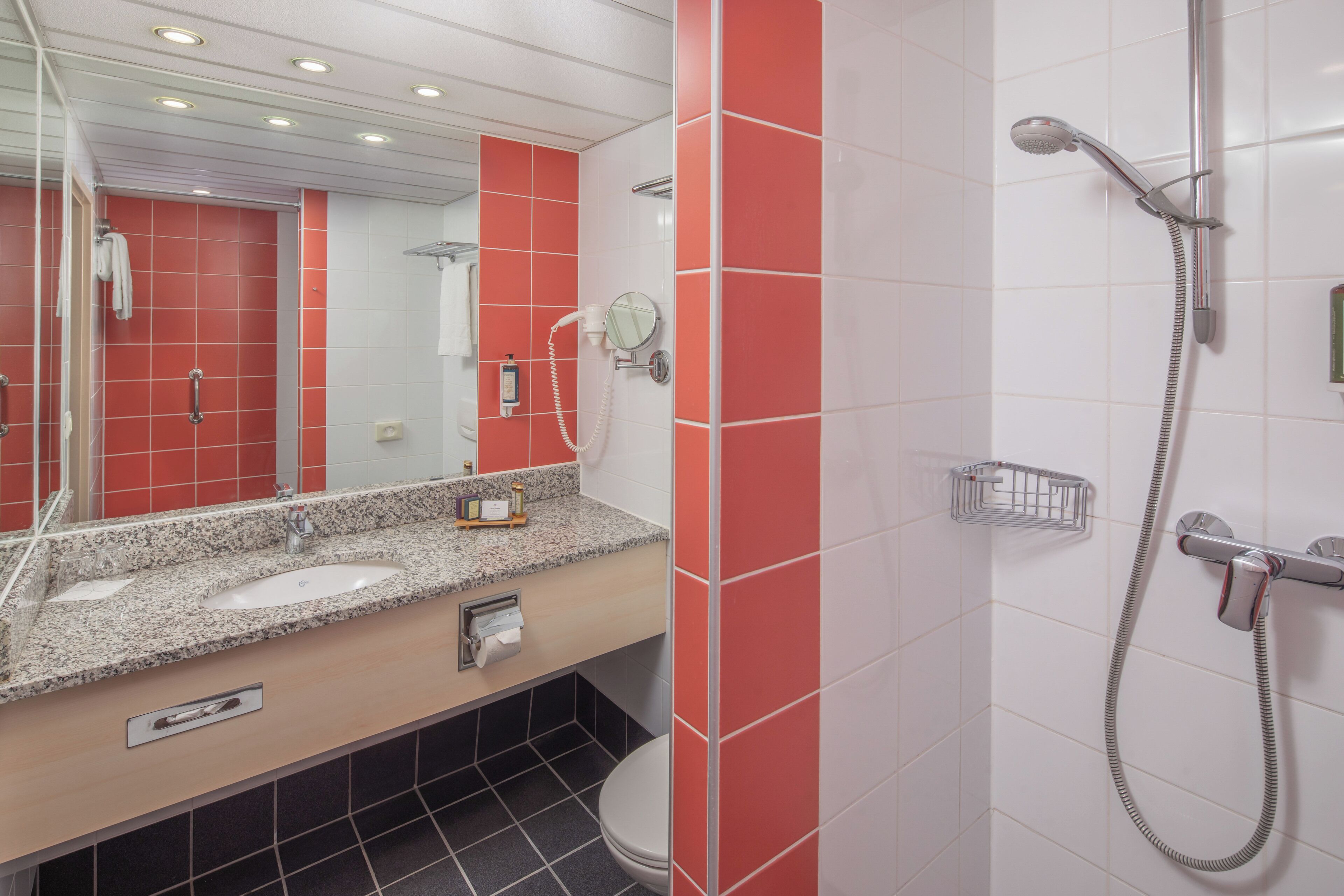 A modern bathroom with a granite countertop sink, a toilet, a shower with a handheld showerhead, and a mirror.